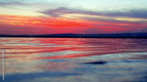 Sunset sky reflects on the calm water surface, painting it in pink, violet, and blue tones. The horizon is lined with distant hill silhouettes