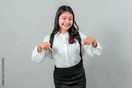 Asian teenage girl in school uniform pointing downward with both hands, smiling confidently. Studio shot on gray background. Back to school concept, education and student life theme.