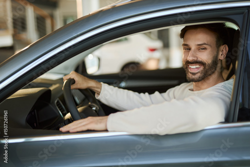 Happy man sitting in his new car at showroom and looking at camera.