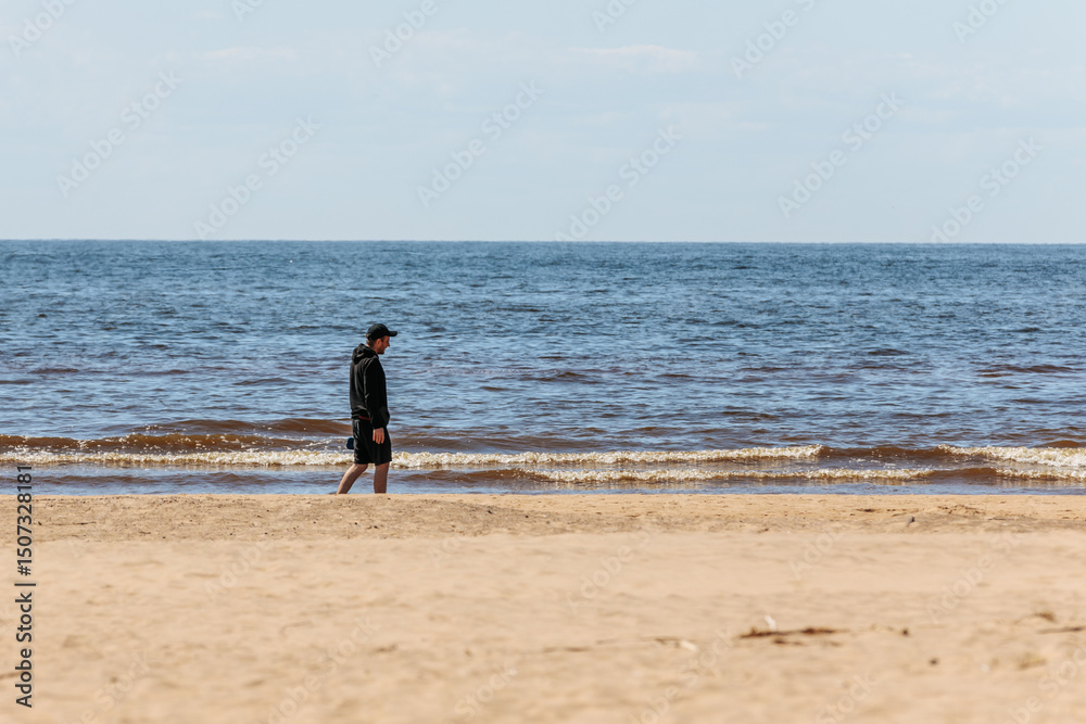A man in a black hoodie and cap walks alone on a sandy beach beside calm waves under a pale blue sky, creating a peaceful, introspective scene. A man in a black hoodie and cap walks alone on a sandy beach beside calm waves under a pale blue sky, creating a peaceful, introspective scene.