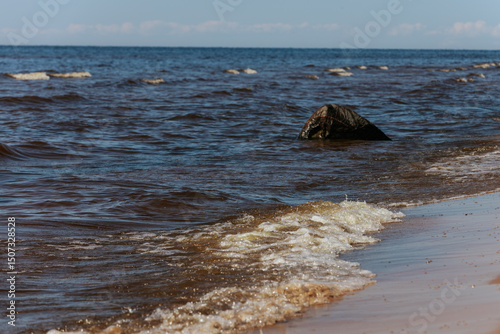 Fototapeta Naklejka Na Ścianę i Meble -  A dark rock partially submerged in sea water with small waves crashing on a sandy beach under sunlight.