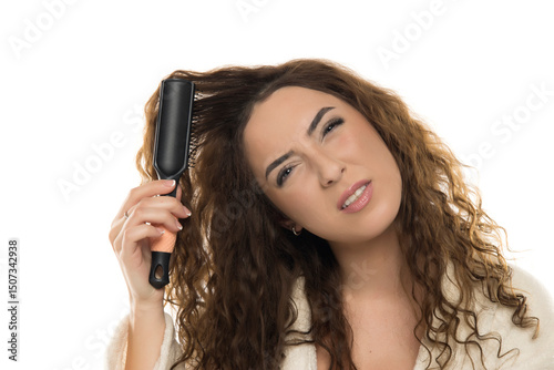 Woman with curly brown hair in white bathrobe struggling with hair brush caught in tangled hair against white background, frustrated expression, styling challenge