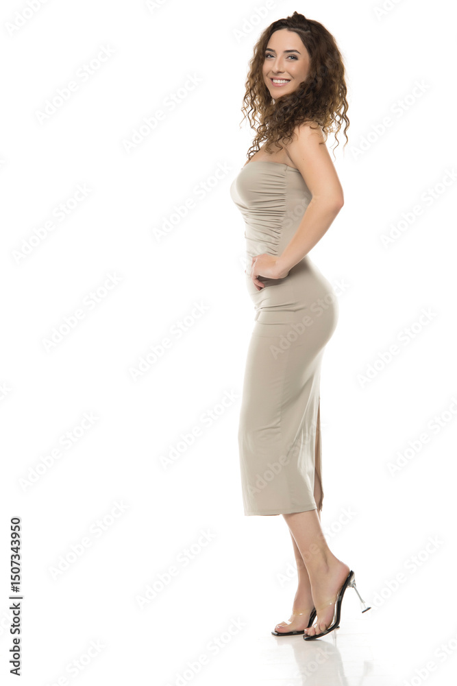 Smiling woman with curly brown hair in beige strapless dress with side slit looking over shoulder against white background, profile fashion portrait pose Smiling woman with curly brown hair in beige strapless dress with side slit looking over shoulder against white background, profile fashion portrait pose