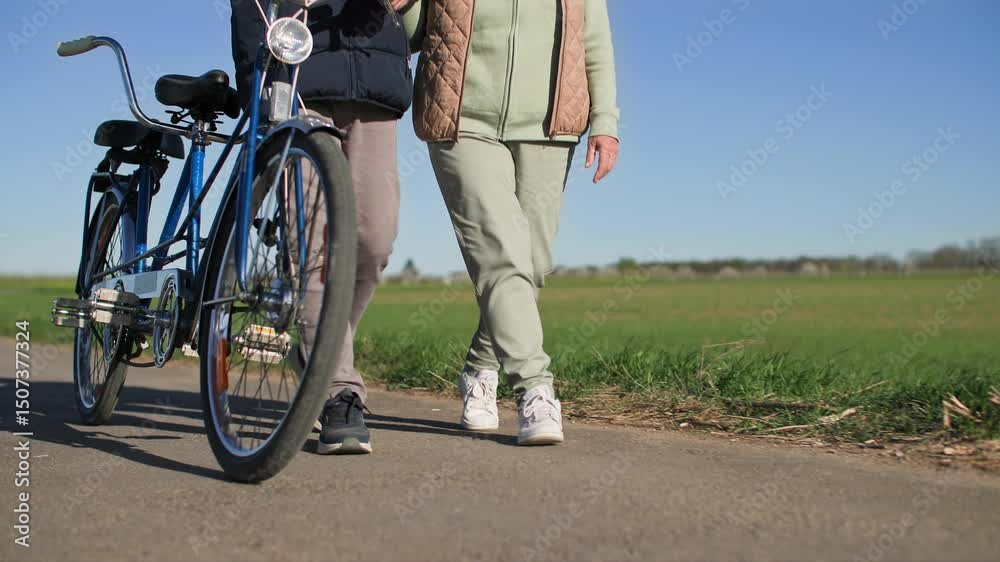 Pension couple with a bicycle walk synchronously along the road past green meadows. View from below on legs. Medium shot. Slow motion