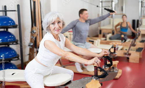 Senior female during Pilates training performs exercises and tasks on heron pulley tower for muscles of arms and body. Non-traumatic sport and fitness, independent and group classes in gym, at home photo