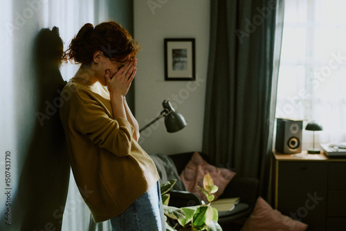 Foto Red-haired woman standing against wall in dimly lit room covering her face, appearing distraught