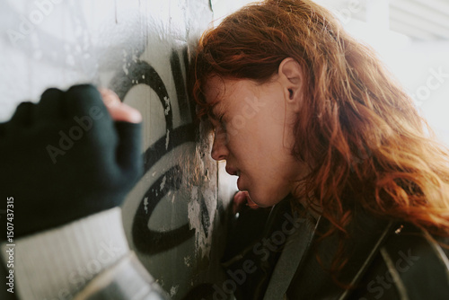 Side-view of woman with red hair leaning against a wall covered in graffiti, captured in depressive moment. Wearing dark jacket, facing away from the camera