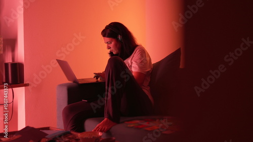 Fotografie Woman working on laptop at home in a dimly lit room, headphones on, sitting on a