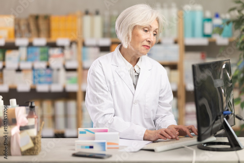 Wallpaper Mural Mature female pharmacist standing behind the counter and working on computer, she checks the availability of medical medicines in a spreadsheet and prepares to collect an online order Torontodigital.ca