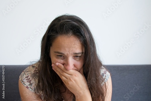 Worried woman sitting on a sofa covering her mouth with her hand, expressing anxiety and concern