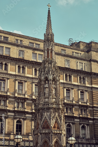 Ornate Eleanor Cross in Charing Cross, London, featuring Gothic architecture with a detailed spire and statue, set against a historic building under a partly cloudy sky.