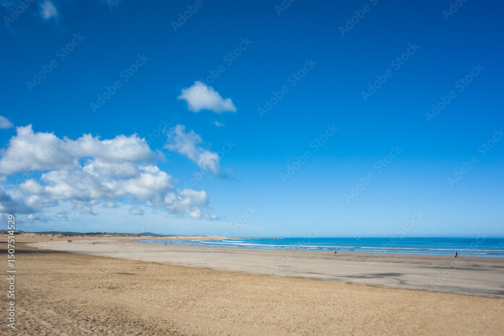 Morning view of the wide sandy beach in Essaouira, Morocco, with low tide, clear blue sky over the sea, fluffy clouds over land, and few distant people walking along the shore