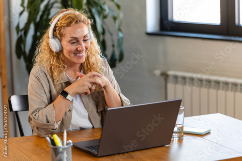 Smiling woman with headphones attentively watching a laptop screen during an online meeting, seated at a wooden desk in a cozy home office.