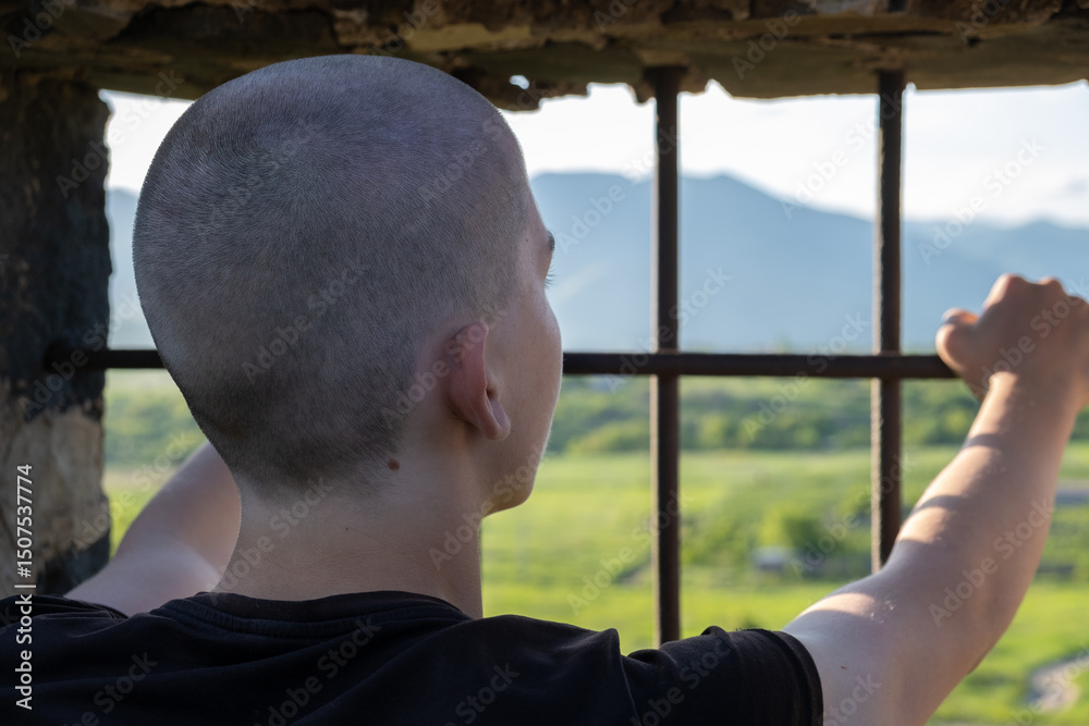 Fototapeta premium Rear view of a shaved-headed teenage boy in a solitary prison cell, leaning on the barred window and gazing at the sunset and mountains beyond. Longing for freedom and missing home.