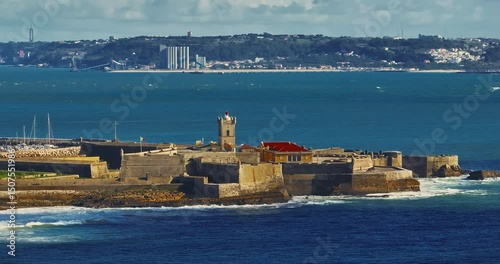 Drone view of a fortress in Portugal with the famous bridge in the background. Saint Julian Fortress with lighthouse tower from beach of Carcavelos, Portugal. Atlantic ocean waves beach in Carcavelos
