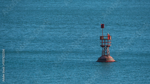 A red navigational buoy stands alone in calm blue ocean waters under natural light, marking a sea channel