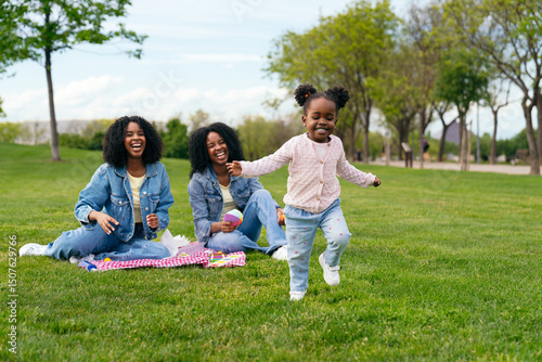 Cheerful toddler running on grass while her mother and aunt are sitting on blanket having picnic in park
