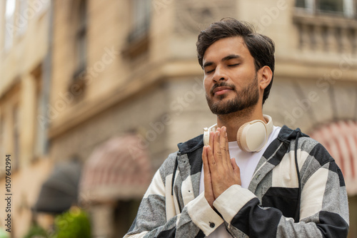 Religious Indian man tourist praying with closed eyes to God asking for blessing, help, forgiveness outdoor. Young guy in casuals clasping hands wishing luck in urban sunny city street. Town lifestyle photo