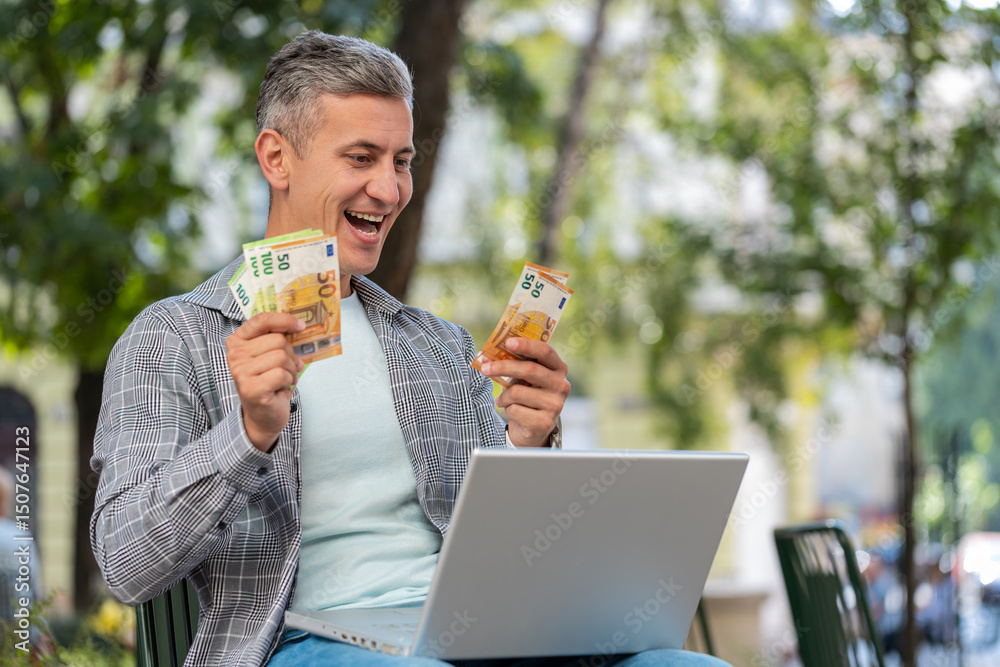 Fototapeta premium Happy rich gray-haired man using laptop winner victory counting holding fan of cash money in euro banknotes sitting on chair city street park. Young male guy tourist planning budget online shopping