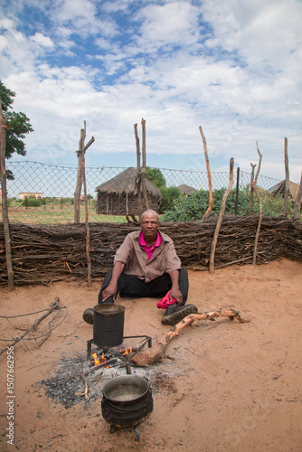 african village senior old african man, in the kitchen cooking on cauldron, in front of house , home with thatched roof, fencing , rural living