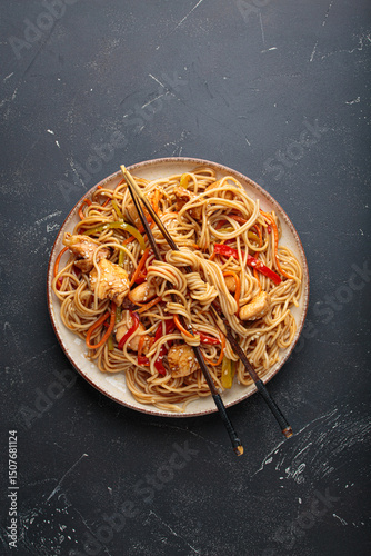 Delicious udon noodles stir fried with chicken meat and vegetables, sprinkled with sesame seeds, served on a plate with chopsticks on a dark background, creating an appetizing asian cuisine scene