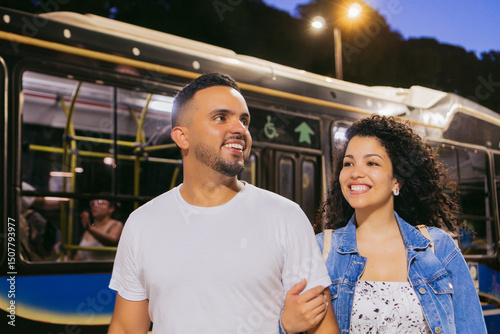 Young Latin woman happily holds her partner's arm to walk after getting off the bus behind them