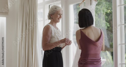 Diverse women smoothing gown, applying powder, showing jewelry during wedding prep at bridal suite