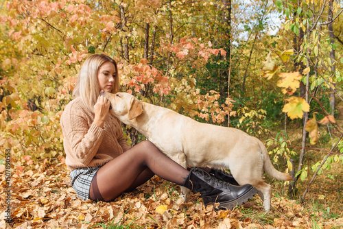 Young beautigul caucasian woman walking with her beloved labrador dog in autumn park. Woman sitting on the ground with fallen golden leaves hugging her dog.