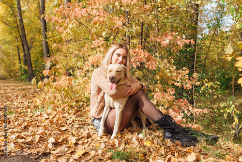 Young beautigul caucasian woman walking with her beloved labrador dog in autumn park. Woman sitting on the ground with fallen golden leaves hugging her dog.