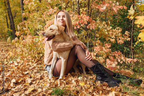 Young beautigul caucasian woman walking with her beloved labrador dog in autumn park. Woman sitting on the ground with fallen golden leaves hugging her dog.