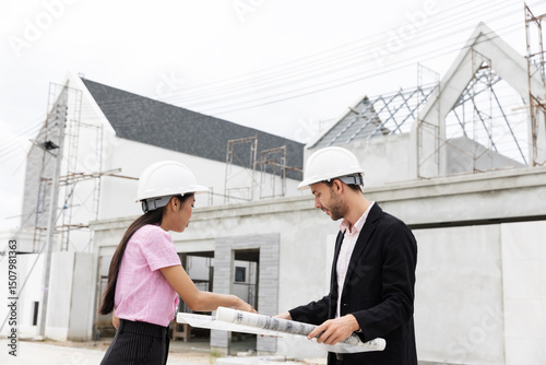 Two architects wearing safety helmets discuss blueprints at a construction site. Surrounded by scaffolding and a partially built structure, they collaborate on the details of the building project.