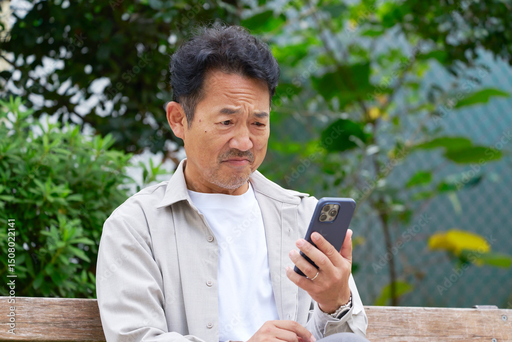 Fototapeta premium 悩まみながら屋外でスマートフォンを見る中年の男性 屋外