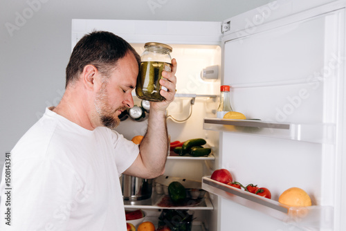 Man holding jar of pickles against forehead, standing in front of open refrigerator filled with fresh vegetables and fruits, conveying the struggle of dealing with headache and hangover symptoms