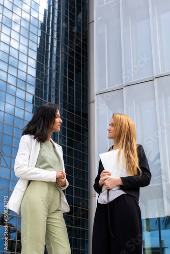 Urban scene with women discussing business strategy outdoors, modern glass architecture, leadership and success concept, elegant and determined expressions