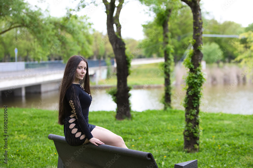 Latin woman, young and beautiful brunette dressed in black sitting on the back of a park bench looks mysteriously and sensually backwards. In the background a lake on a gray day. Latin woman, young and beautiful brunette dressed in black sitting on the back of a park bench looks mysteriously and sensually backwards. In the background a lake on a gray day.