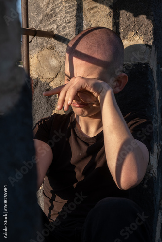 Canvas Print Vertical photo of a shaved-head teenage boy in a prison cell, sitting by a barred window and wiping tears from his face