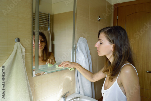 Person photographed while cleaning the house: a woman wipes a glass shelf under a mirror in the bathroom with a cloth. Keeping your apartment clean by yourself: saving on cleaning services.