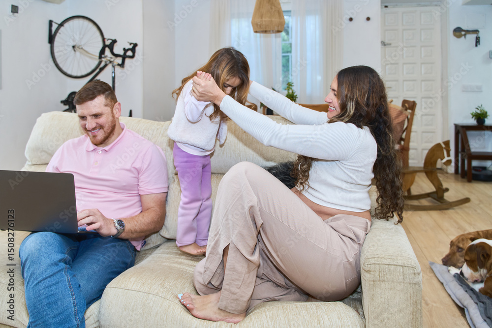 Fototapeta premium Pregnant mother playing with daughter while father works on laptop