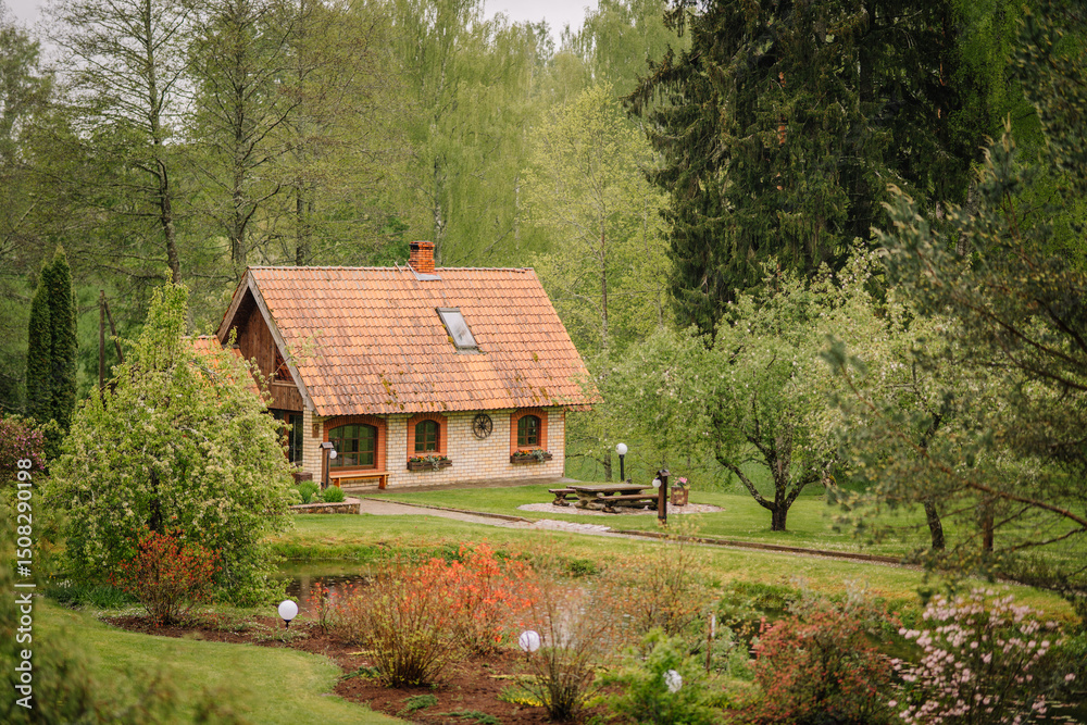 Obraz premium Charming brick cottage with tiled roof in a lush green garden, surrounded by trees, flowers, and a pond with springtime blooms..
