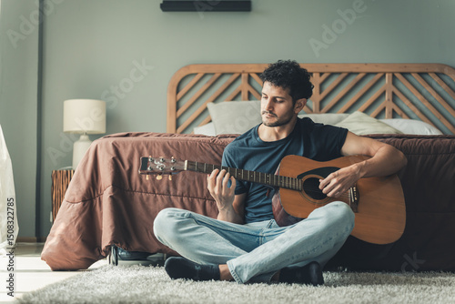 Male guitarist sitting on carpeted floor with acoustic guitar across legs, practicing quietly in relaxed bedroom space, portraying peaceful creativity and passion for music, man on holiday at home