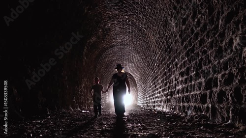 A woman and a child walk out of a dark tunnel into the light, casting dramatic silhouettes against the lit stone wall. A symbolic shot of transition, protection, and generational bond.