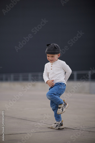 Cute little boy wearing a stylish cat-eared hat, white shirt, and jeans, walking confidently on an urban sidewalk with a concrete and metal background. Casual outdoor children’s fashion.