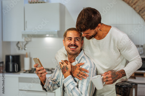Tattooed gay couple hugging while drinking coffee and using smart phone