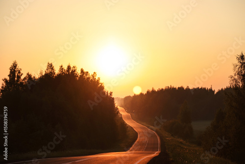 Bright sun at sunrise over the road and forest. Morning summer landscape at dawn