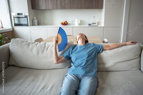 Woman lounging on sofa fanning herself in hot airless room without air conditioning. Elderly discomfort from sweltering, heatwave vulnerability, poor ventilation struggle, aging female hardship photo