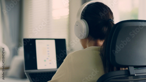 Back view of Asian woman wearing headphones while working on laptop at home. Remote work, online meeting, or digital focus in modern indoor workspace with natural light.