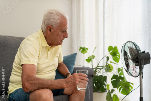Senior man suffering from summer heatwave holds glass of water while sitting on the sofa near a fan