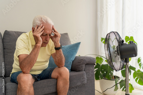 Konstfotografi Senior man suffering from heat while sitting at home in front of fan holding hea