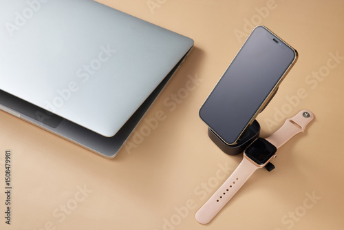 Laptop with smart watch and smartphone charging on wireless charger on beige table, Modern workplace device