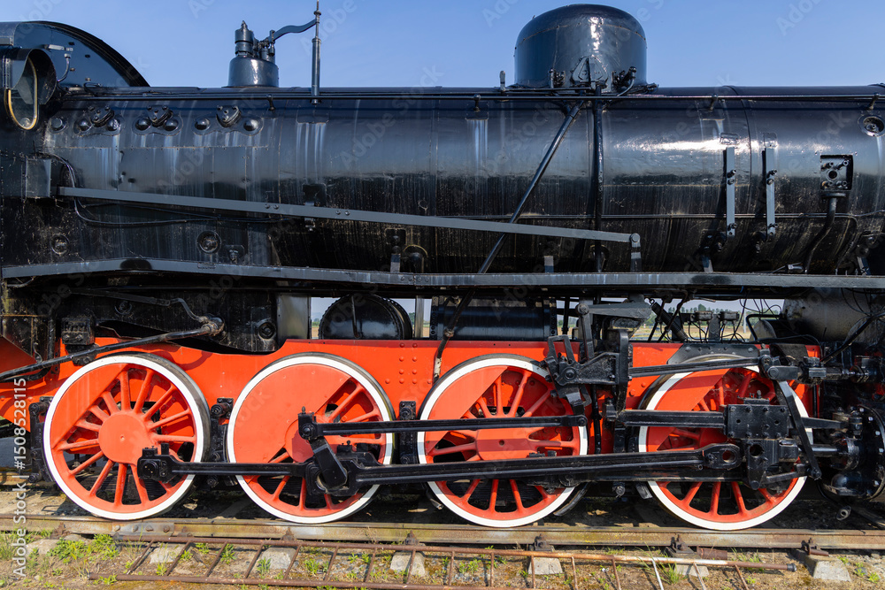 Naklejka premium Old black steam locomotive with red wheels standing on rails against blue sky